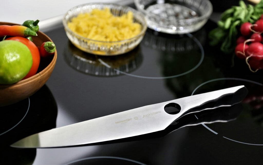 "Close-up of a modern stainless steel kitchen knife placed on a sleek black induction cooktop, surrounded by fresh ingredients including a wooden bowl of limes, tomatoes, chili peppers, and radishes, with a bowl of uncooked pasta in the background."