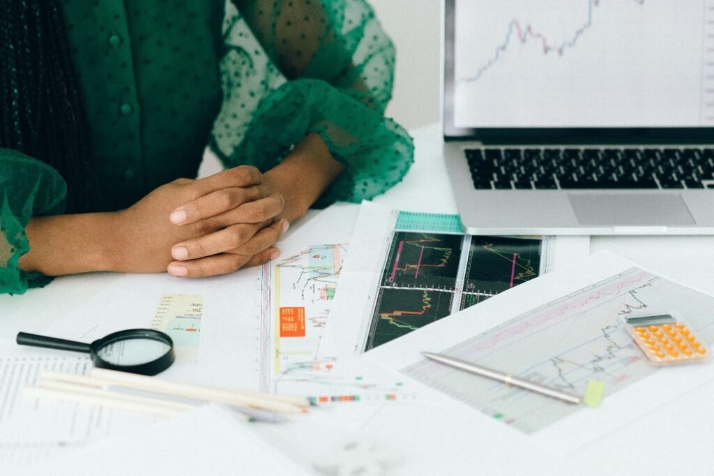 A person wearing a green polka-dot blouse sits at a desk with financial charts, graphs, and a laptop displaying a rising stock market graph. A magnifying glass, calculator, pen, and ruler are also present, suggesting financial analysis or stock market research.