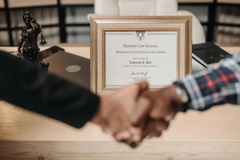 A close-up of two individuals shaking hands in an office setting. In the background, a framed certificate from Harvard Law School is displayed on a wooden desk, along with a small statue of Lady Justice, a laptop, and some books. The image conveys professionalism, trust, and legal expertise.