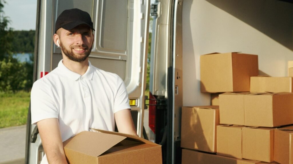 A delivery worker in a white polo shirt and black cap is smiling while holding a cardboard box in front of an open van filled with packages.