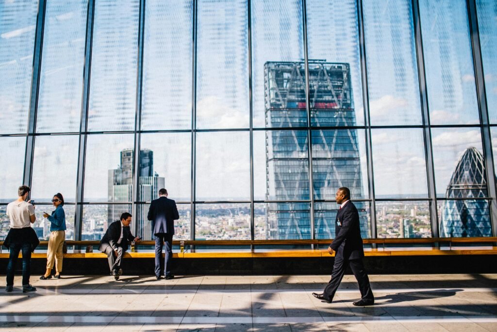 A spacious, modern indoor area with a large glass wall offering a panoramic view of a city skyline, including skyscrapers. Several professionally dressed individuals are engaged in various activities—some standing, walking, or sitting on a wooden bench, while others converse or look at their phones. The reflections on the glass add depth to the scene, capturing a dynamic yet professional atmosphere.